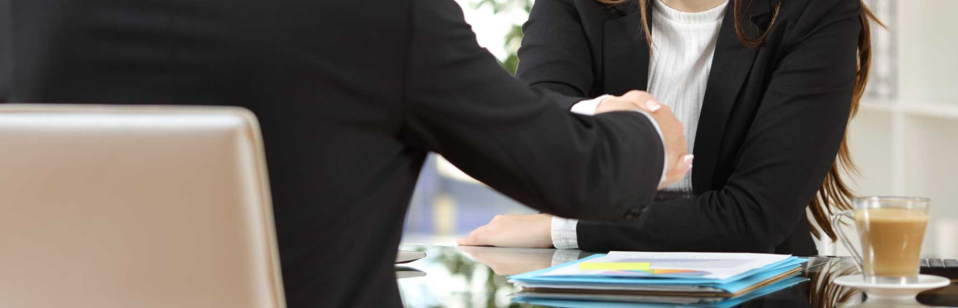 Two people shaking hands across a desk at the end of a workplace meeting